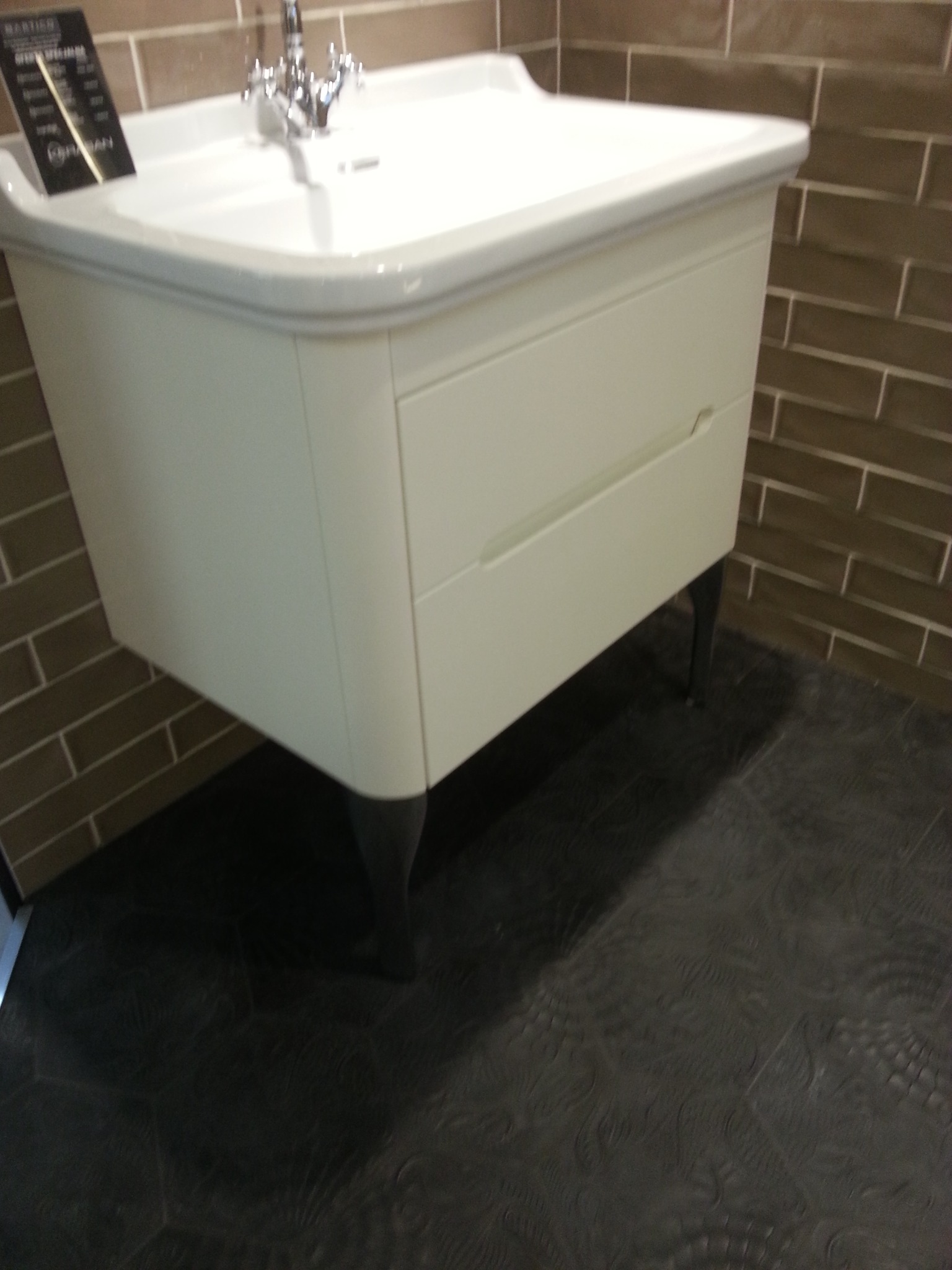 Biała szafka łazienkowa z umywalką i chromowaną baterią na tle brązowych płytek imitujących cegłę. Widoczna podłoga z ciemnej, wzorzystej terakoty.