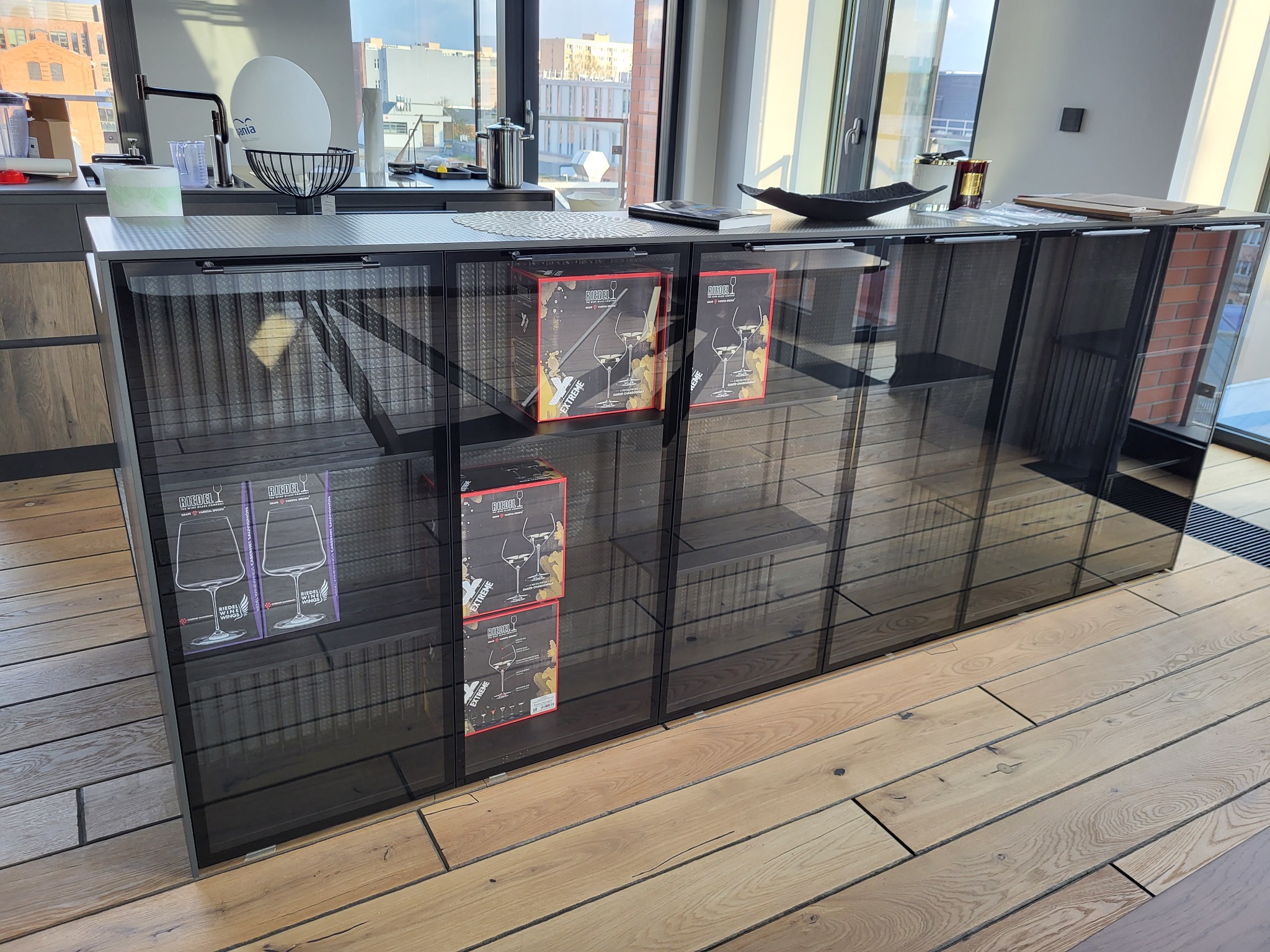 Szklana witryna kuchenna z szufladami, na drewnianej podłodze, z pudełkami kieliszków Riedel w środku. W tle widok na miasto przez okno.