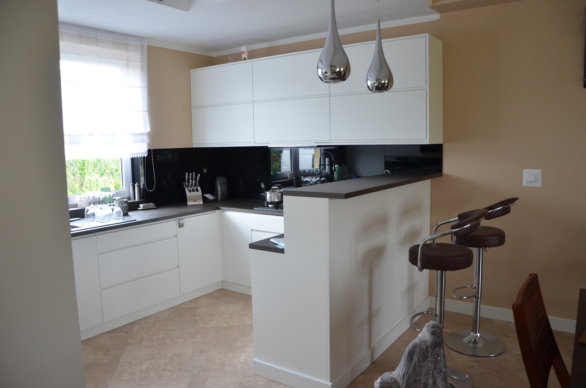 Nowoczesna kuchnia z białymi szafkami, czarnym panelem szklanym i barkiem z hokerami. Dwa srebrne, wiszące światła nad blatem. Jasna podłoga i ściany.