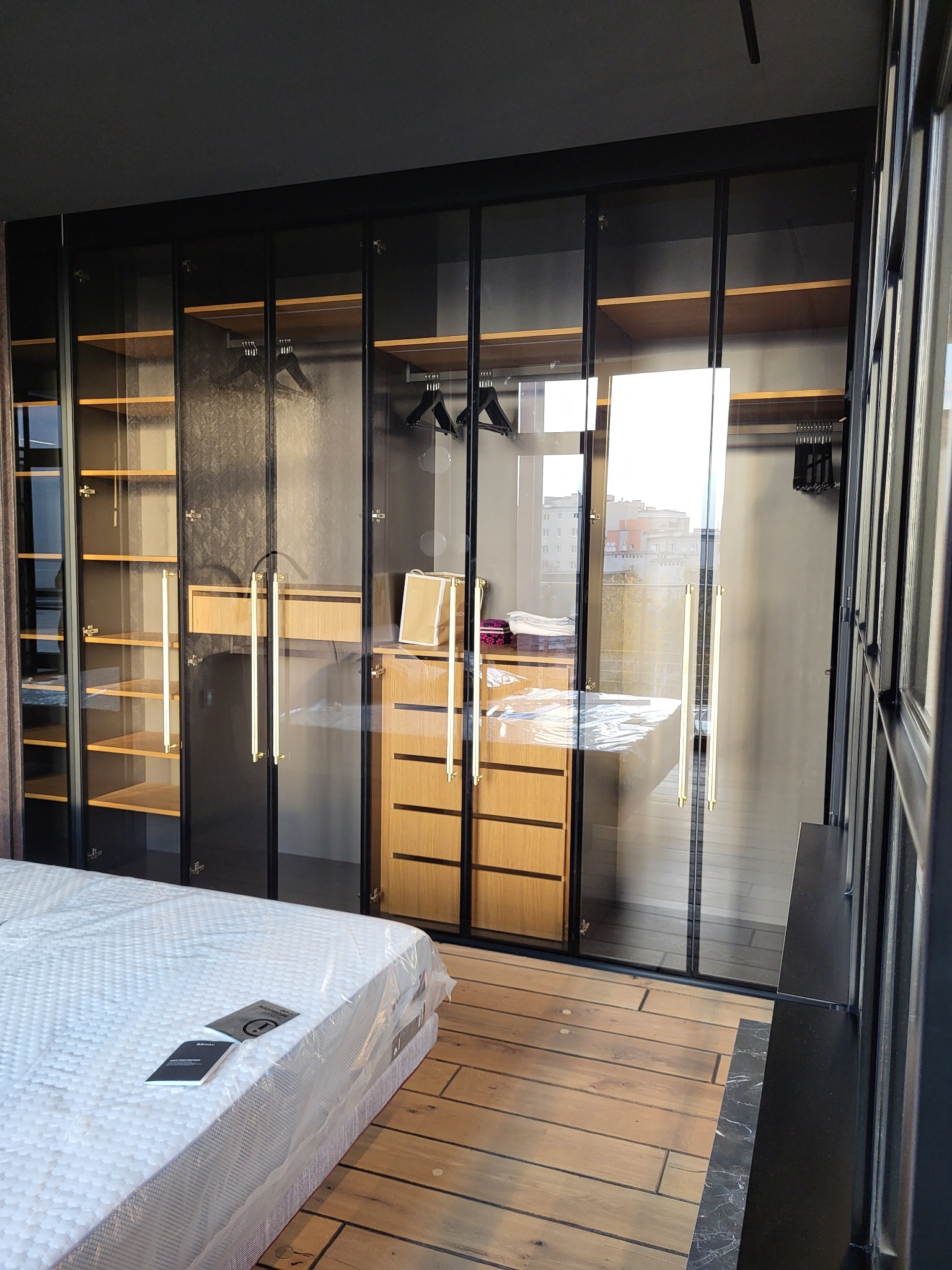Nowoczesna szafa wnękowa z przesuwnymi, szklanymi drzwiami w czarnej ramie. Widoczne półki i szuflady z jasnego drewna. Podłoga z drewnianych paneli.