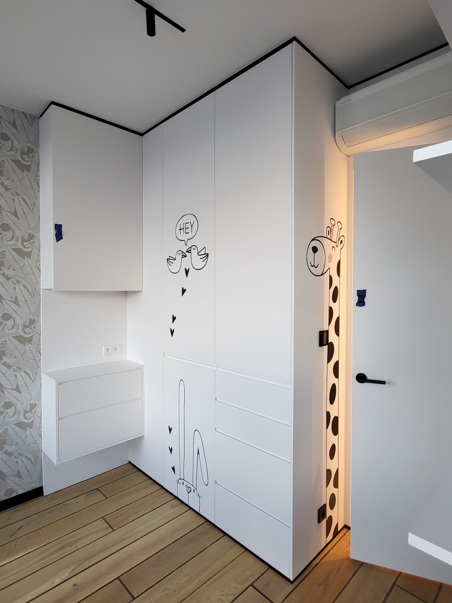Biała zabudowa meblowa w pokoju dziecięcym z rysunkami zwierząt: żyrafy, króliczka i ptaków. Minimalistyczny design z drewnianą podłogą i czarnymi akcentami.