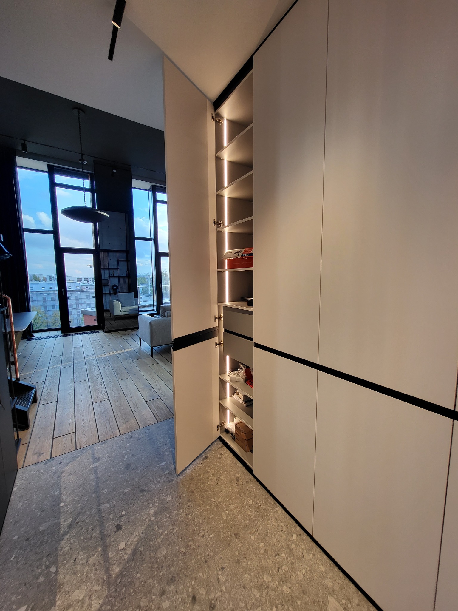 Nowoczesna szafa wnękowa z oświetleniem LED, widok na wnętrze z półkami i szufladami. W tle salon z dużymi oknami i drewnianą podłogą.