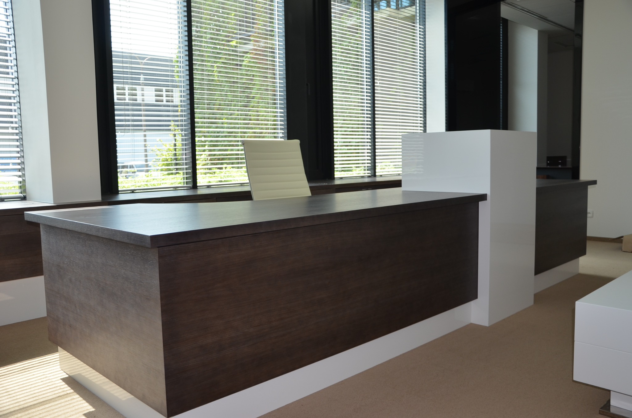 Nowoczesna zabudowa biurowa z ciemnego drewna i białych elementów. Krzesło biurowe stoi przy blacie na tle okna z żaluzjami. Jasna podłoga.