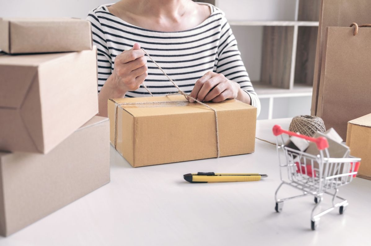 Osoba wiąże sznurkiem kartonowe pudełko na białym stole. Obok leżą inne pudełka, nożyk i miniaturowy wózek sklepowy. W tle widoczne półki.