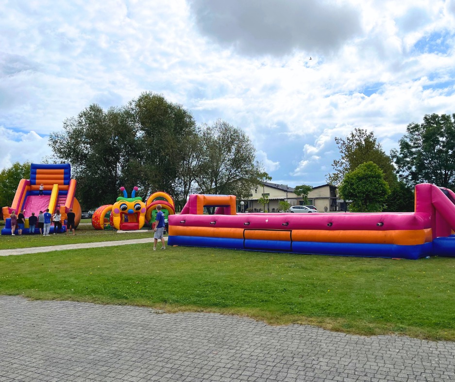 Kolorowe dmuchańce na zielonym trawniku, w tle drzewa i budynek. Widoczny dmuchany zamek ze zjeżdżalnią, tunel w kształcie gąsienicy i tor przeszkód.