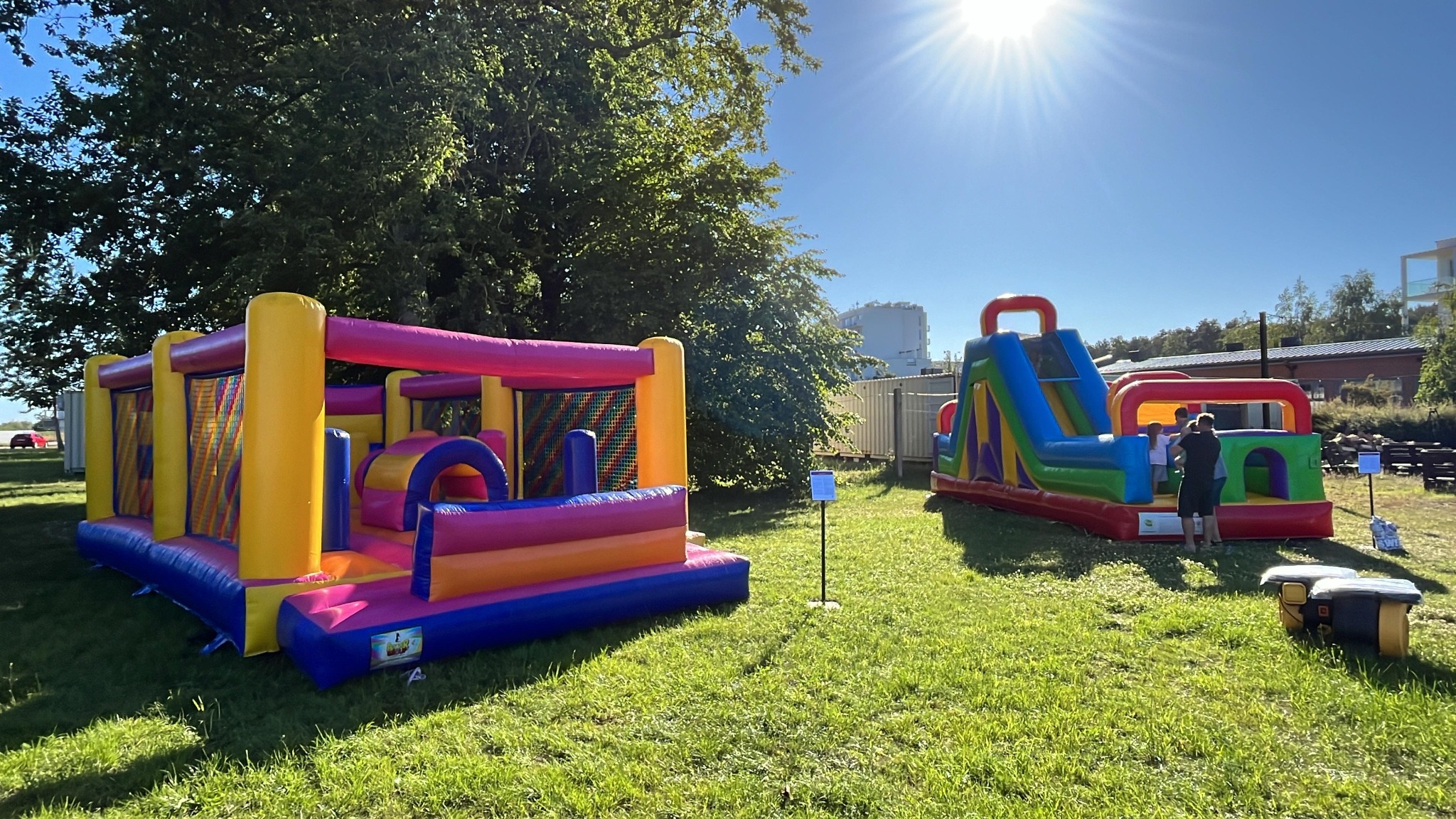 Dwa kolorowe dmuchańce na trawie w słoneczny dzień. Jeden z nich to zamek, drugi ze zjeżdżalnią. W tle drzewa i budynek.
