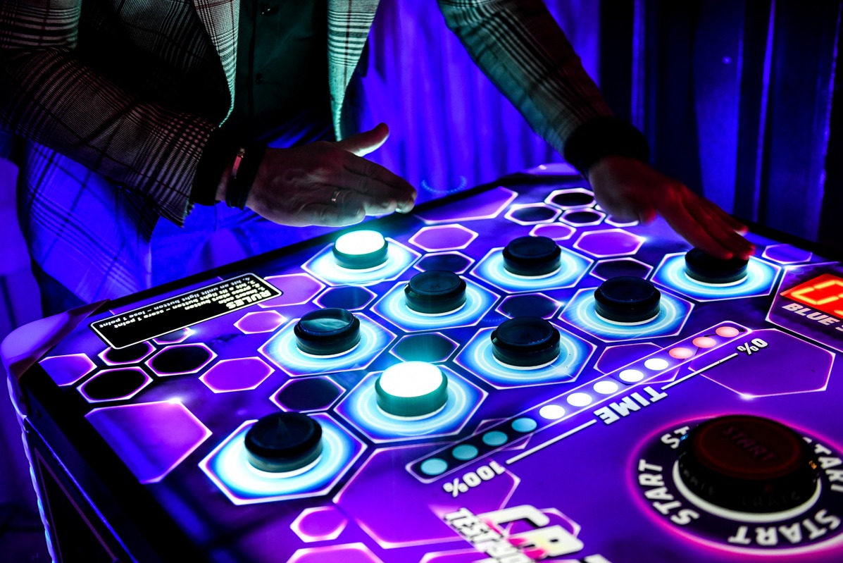 Interaktywny stół z podświetlanymi przyciskami w kształcie heksagonów, obsługiwany przez osobę w marynarce w kratę, w neonowym oświetleniu. Widoczny panel sterowania.