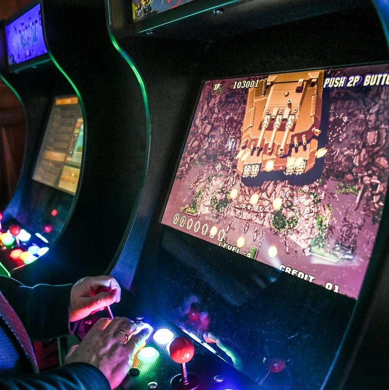 Automat do gier arcade z gra wojenną na ekranie, oświetlony neonowymi przyciskami, obsługiwany przez dłoń gracza z pierścieniem, w tle drugi automat.