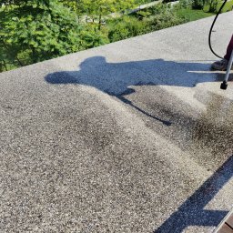 ALPSOLUTIONS SPÓŁKA Z OGRANICZONĄ ODPOWIEDZIALNOŚCIĄ - Czyszczenie dachu z kamyczkową fakturą za pomocą myjki ciśnieniowej. Widoczny cień osoby wykonującej prace i fragment mokrej powierzchni po czyszczeniu.