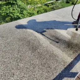 Czyszczenie dachu z kamyczkową fakturą za pomocą myjki ciśnieniowej. Widoczny cień osoby wykonującej prace i fragment mokrej powierzchni po czyszczeniu.