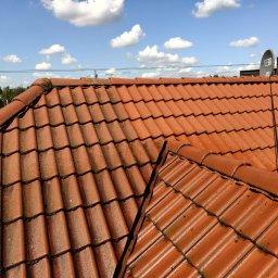 ALPSOLUTIONS SPÓŁKA Z OGRANICZONĄ ODPOWIEDZIALNOŚCIĄ - Czerwona dachówka z widocznymi śladami mchu i zabrudzeń, na tle błękitnego nieba z białymi obłokami. Widoczne anteny satelitarne i fragmenty innych budynków.