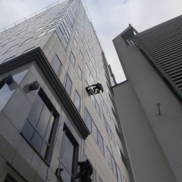 ALPSOLUTIONS SPÓŁKA Z OGRANICZONĄ ODPOWIEDZIALNOŚCIĄ - Pracownicy wysokościowi na linach czyszczą okna biurowca z szarej elewacji. Widok z dołu na budynek i pochmurne niebo.