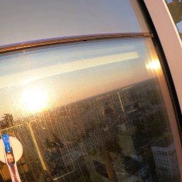 ALPSOLUTIONS SPÓŁKA Z OGRANICZONĄ ODPOWIEDZIALNOŚCIĄ - Mycie okien na wysokości w wieżowcu. Widoczna panorama miasta odbijająca się w szybach i narzędzie pracy - myjka do okien z długim uchwytem.