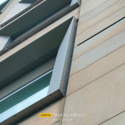 ALPSOLUTIONS SPÓŁKA Z OGRANICZONĄ ODPOWIEDZIALNOŚCIĄ - Niski kąt na elewację z oknem i balkonem. Widoczne zabrudzenia, potencjalnie przed czyszczeniem. Geometryczny układ płyt elewacyjnych.