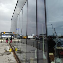 ALPSOLUTIONS SPÓŁKA Z OGRANICZONĄ ODPOWIEDZIALNOŚCIĄ - Dwie osoby myją szklaną elewację budynku komercyjnego, używając ściągaczek i drabin. Widoczne odbicia otoczenia w czyszczonych oknach.