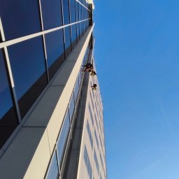 ALPSOLUTIONS SPÓŁKA Z OGRANICZONĄ ODPOWIEDZIALNOŚCIĄ - Dwóch alpinistów w uprzężach czyści okna wieżowca na wysokości, widok z dołu, błękitne niebo w tle.