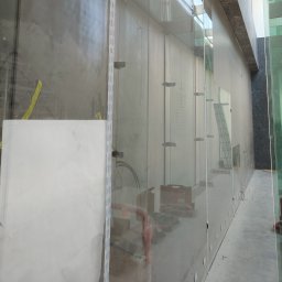 ALPSOLUTIONS Sp. z o.o. - Szklana elewacja budynku w trakcie montażu. Widoczne narzędzia, drabina i elementy mocujące szkło. Surowy beton w tle podkreśla etap prac.