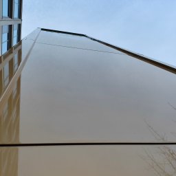 ALPSOLUTIONS SPÓŁKA Z OGRANICZONĄ ODPOWIEDZIALNOŚCIĄ - Błyszcząca, beżowa elewacja budynku biurowego z widocznym odbiciem sąsiedniego budynku i gałęzi drzew na tle błękitnego nieba. Ujęcie z dołu.