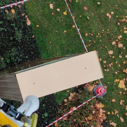 ALPSOLUTIONS Sp. z o.o. - Widok z góry na zabezpieczony obszar robót wysokościowych z użyciem taśmy ostrzegawczej i pachołków, z widocznym fragmentem dachu i zielenią.