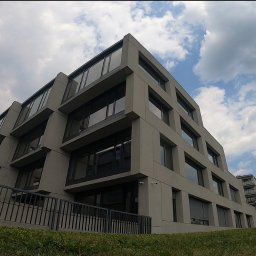 ALPSOLUTIONS SPÓŁKA Z OGRANICZONĄ ODPOWIEDZIALNOŚCIĄ - Nowoczesny, szary budynek biurowy z dużymi oknami, widziany z perspektywy żabiej z trawnika. Na elewacji widoczna kamera monitoringu.