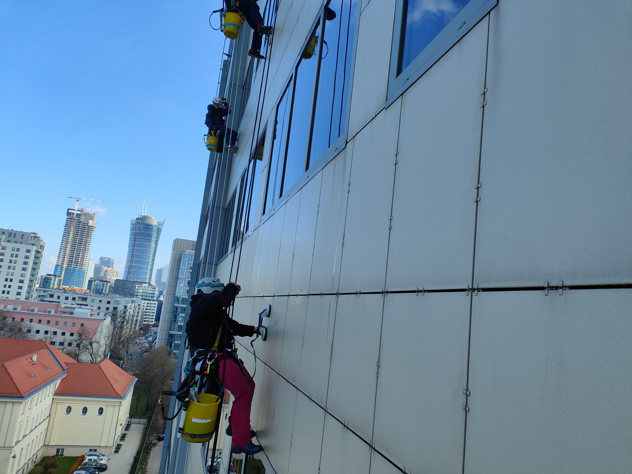 Pracownicy na linach alpinistycznych czyszczą elewację wysokiego budynku biurowego. Widok na panoramę miasta w tle, błękitne niebo. Używają specjalistycznych narzędzi.