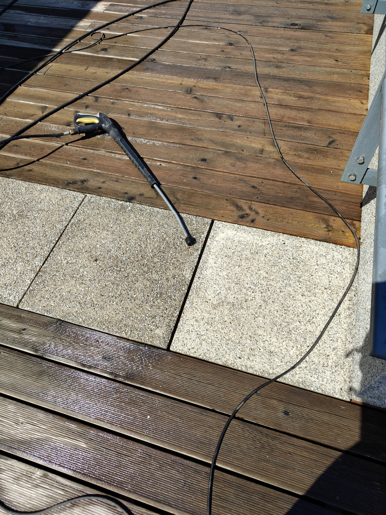 Mycie tarasu z desek i kostki. Częściowo mokra powierzchnia, pistolet do myjki ciśnieniowej i wąż na drewnianym tarasie z elementami kostki brukowej.