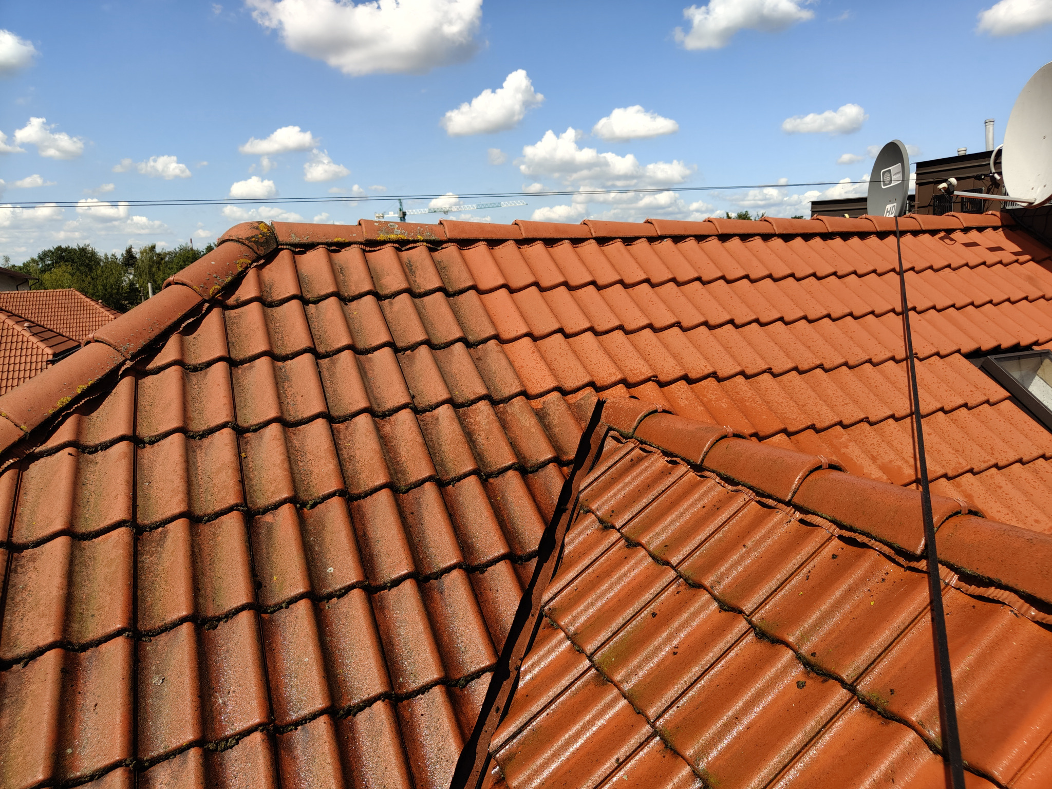 Czerwona dachówka z widocznymi śladami mchu i zabrudzeń, na tle błękitnego nieba z białymi obłokami. Widoczne anteny satelitarne i fragmenty innych budynków.