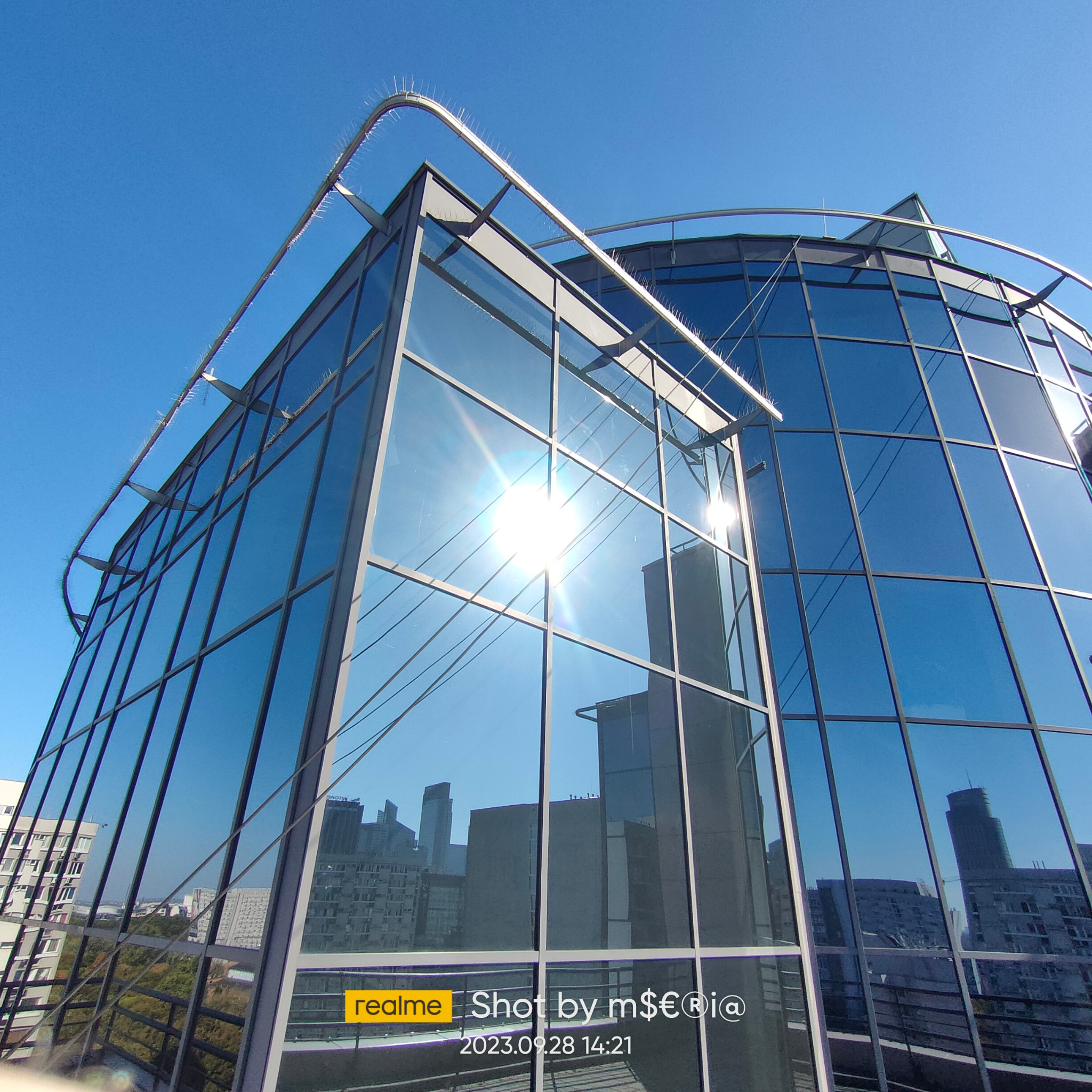 Szklana fasada biurowca z widocznymi kolcami przeciw ptakom na dachu, odbijająca miejski krajobraz w błękitnych szybach.