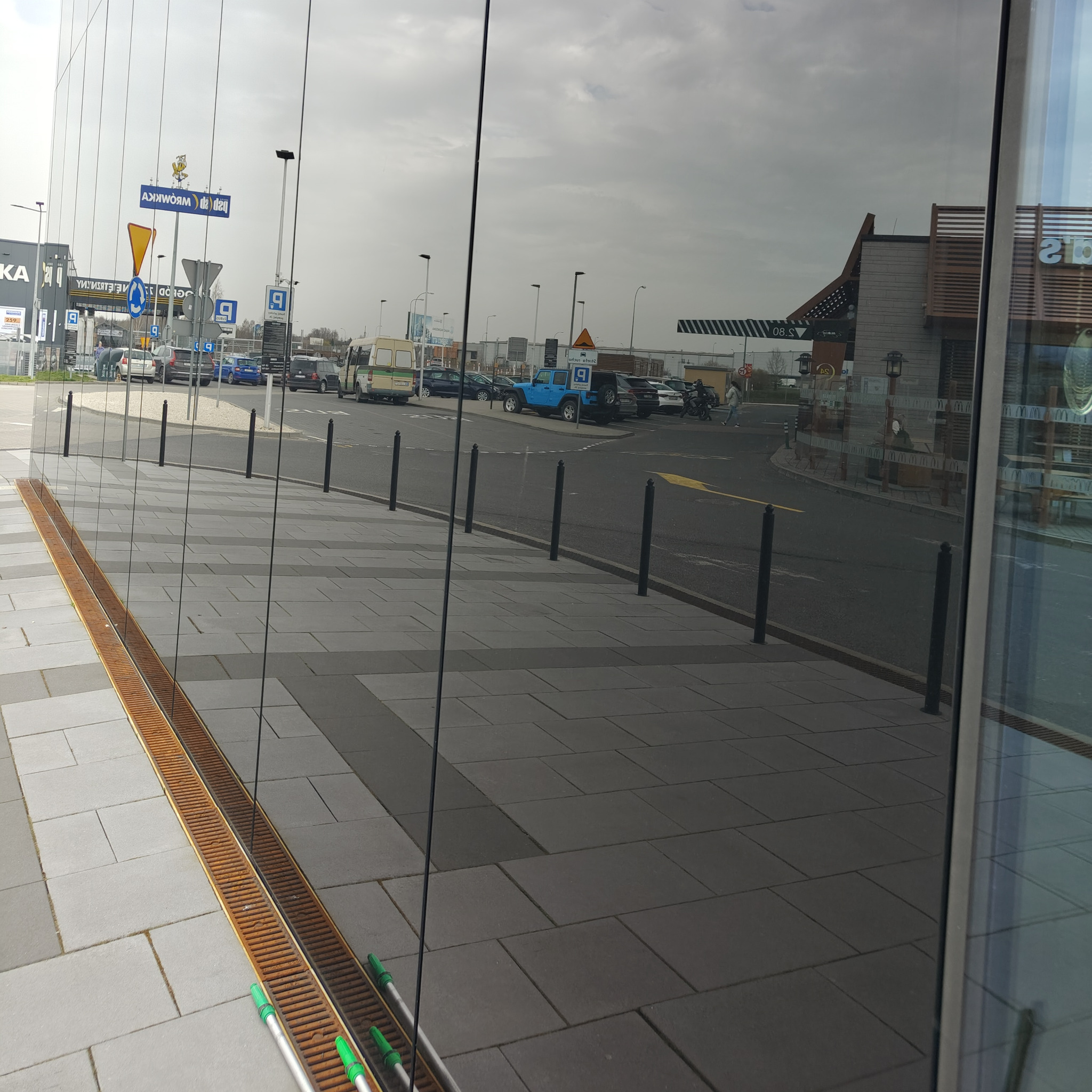 Refleksy na szklanej fasadzie biurowca odbijają parking i ulicę. Widoczne narzędzia do mycia okien. Metalowa kratka odpływowa wzdłuż chodnika.