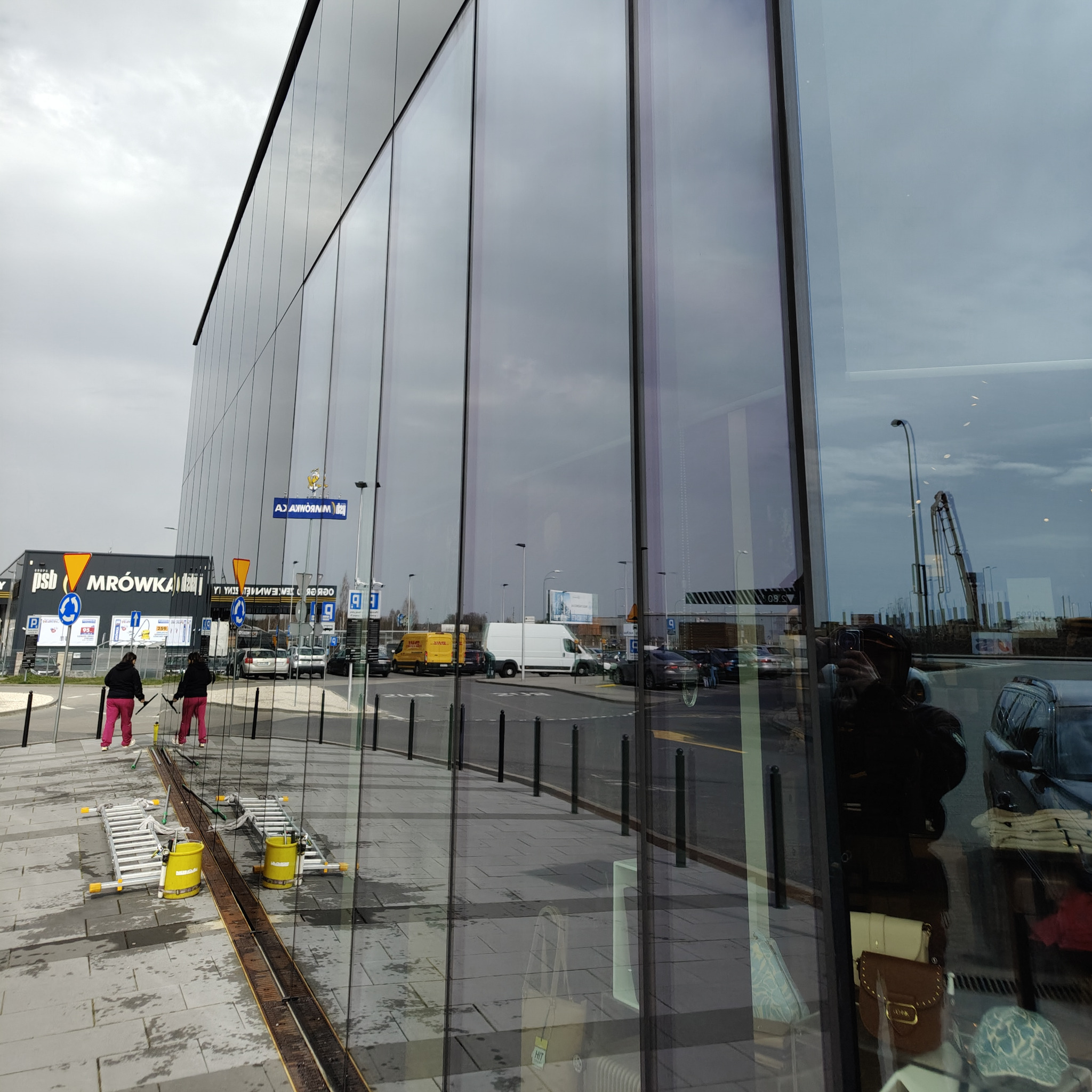 Dwie osoby myją szklaną elewację budynku komercyjnego, używając ściągaczek i drabin. Widoczne odbicia otoczenia w czyszczonych oknach.