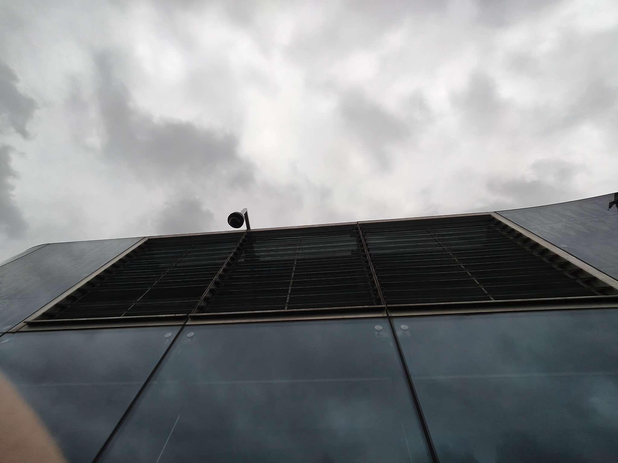 Szklana elewacja budynku z żaluzjami i kamerą na tle pochmurnego nieba, widziana z dołu. Elementy architektoniczne i nowoczesny design.