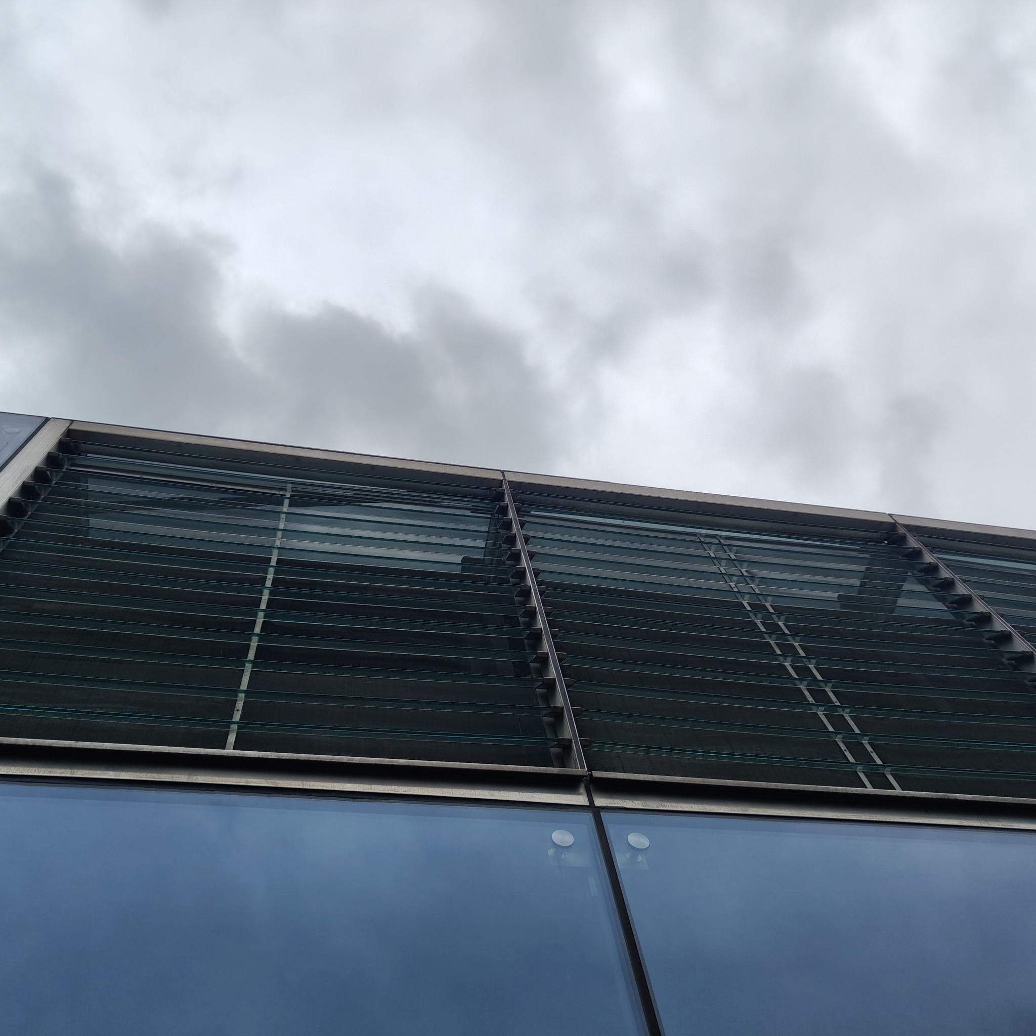 Szklana fasada budynku z żaluzjami zewnętrznymi, widok od dołu w pochmurny dzień. Detal architektoniczny z nowoczesnym designem i stalowymi elementami.