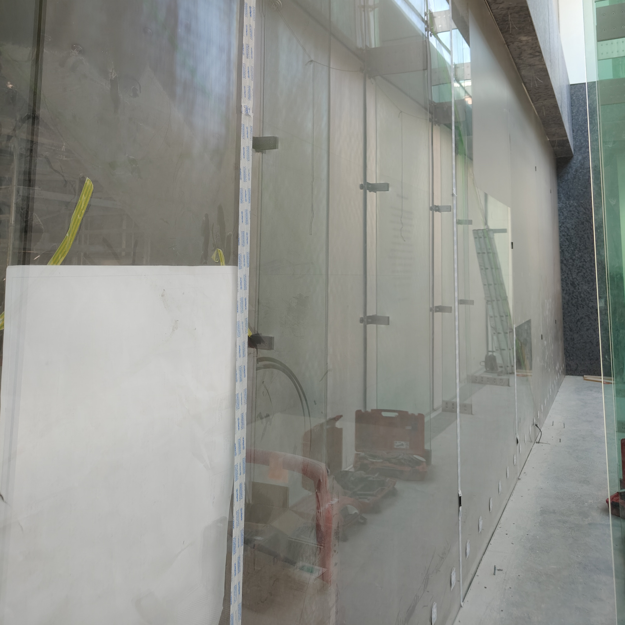 Szklana elewacja budynku w trakcie montażu. Widoczne narzędzia, drabina i elementy mocujące szkło. Surowy beton w tle podkreśla etap prac.