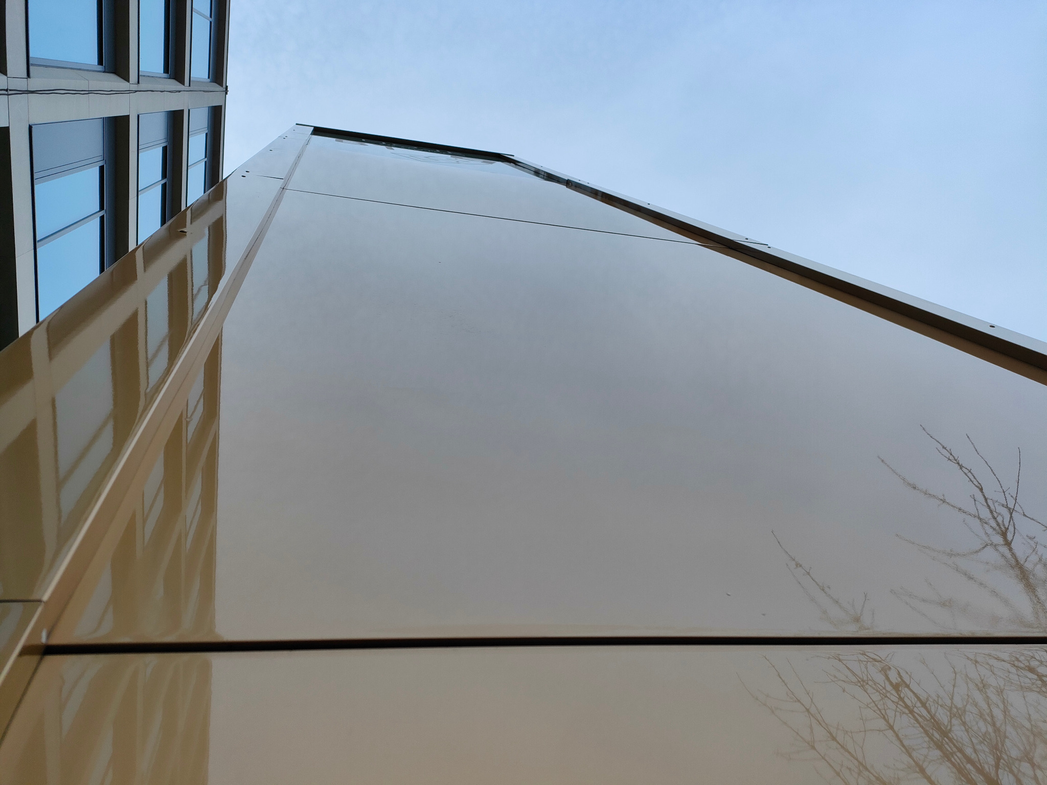 Błyszcząca, beżowa elewacja budynku biurowego z widocznym odbiciem sąsiedniego budynku i gałęzi drzew na tle błękitnego nieba. Ujęcie z dołu.