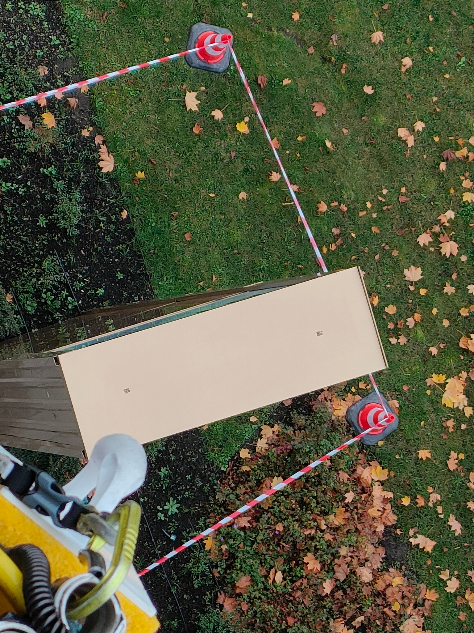 Widok z góry na zabezpieczony obszar robót wysokościowych z użyciem taśmy ostrzegawczej i pachołków, z widocznym fragmentem dachu i zielenią.