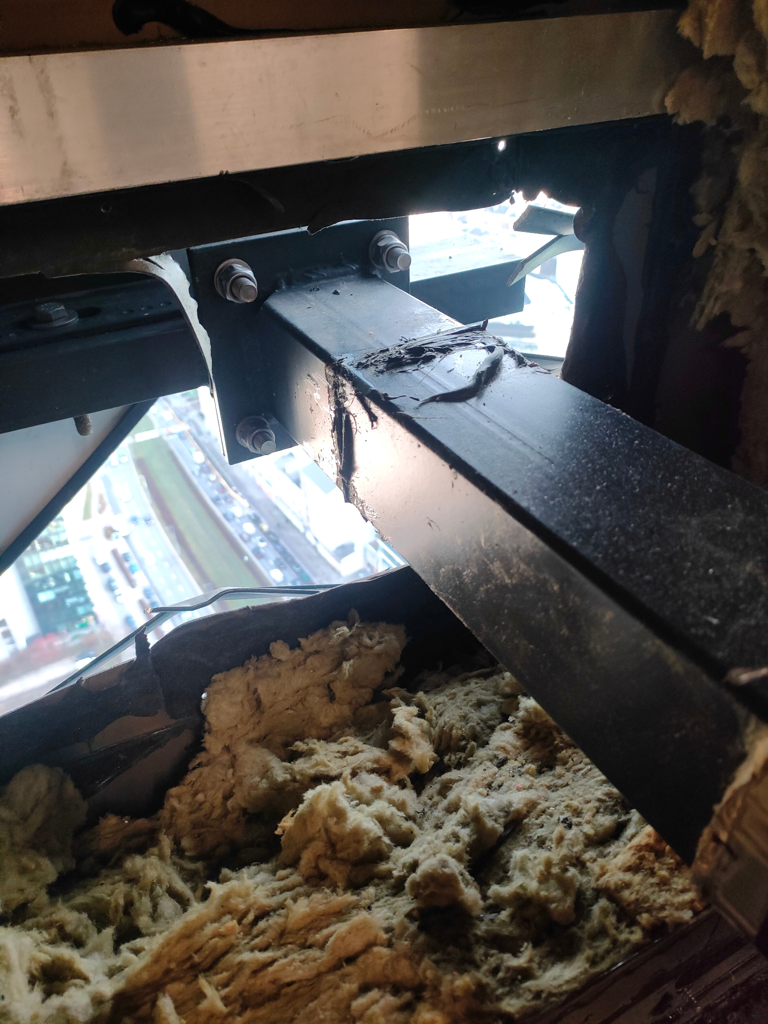 Nietypowy widok zza uszkodzonej konstrukcji okiennej z widoczną izolacją i elementami metalowymi na tle panoramy miasta.