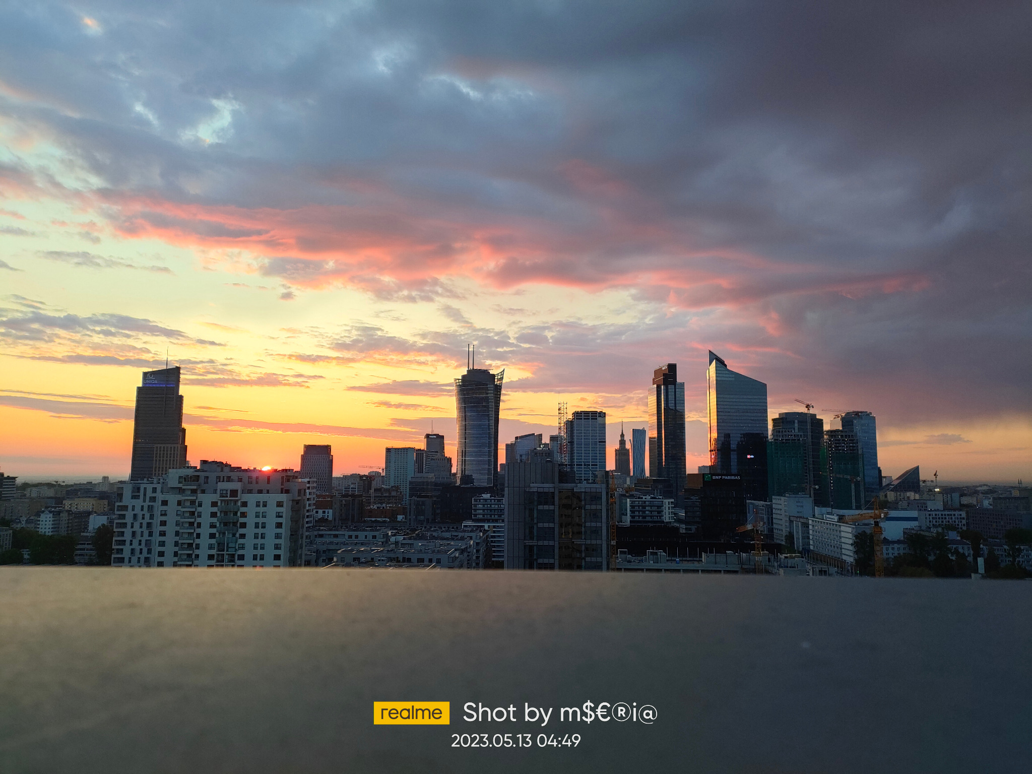 Panorama Warszawy o wschodzie słońca z widokiem na drapacze chmur. Widoczne budynki mieszkalne, biurowce i kolorowe chmury na niebie. Ujęcie z balkonu.