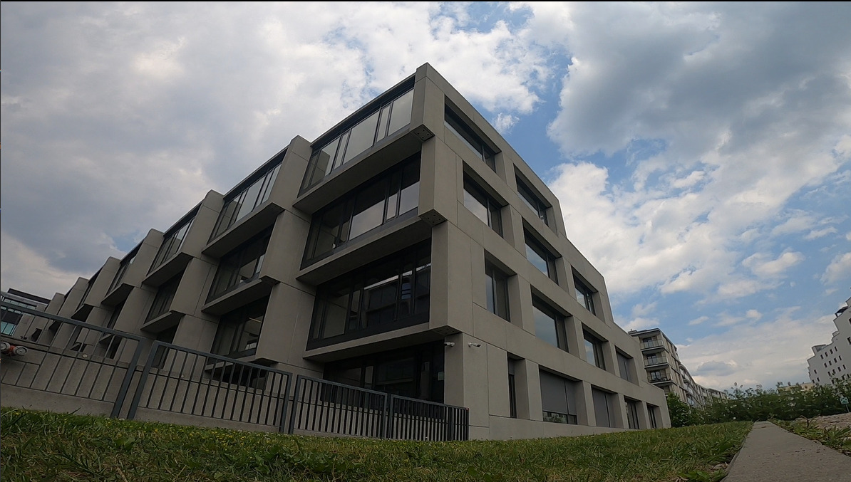 Nowoczesny, szary budynek biurowy z dużymi oknami, widziany z perspektywy żabiej z trawnika. Na elewacji widoczna kamera monitoringu.