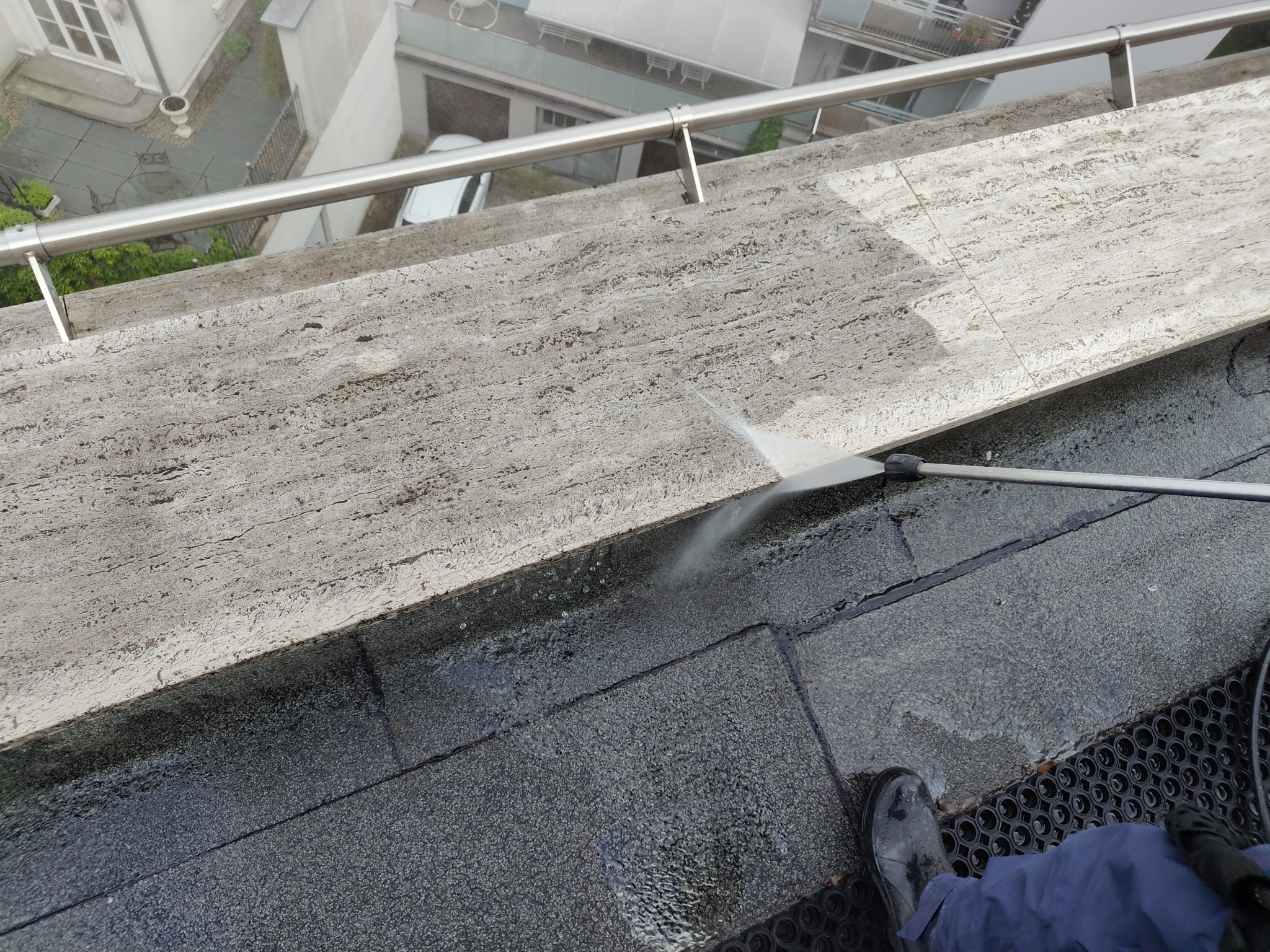 Wysokociśnieniowe czyszczenie krawędzi dachu z marmurowym wykończeniem. Woda spływa po czarnej, gumowej powierzchni dachu. Widoczny fragment buta pracownika.