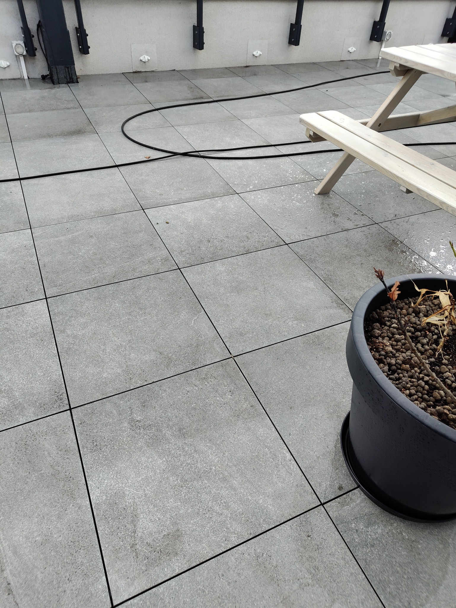 Szare płytki brukowe tarasu po myciu, z widocznymi kroplami wody, czarnym wężem i fragmentem drewnianego stołu piknikowego oraz donicą z suchą rośliną.