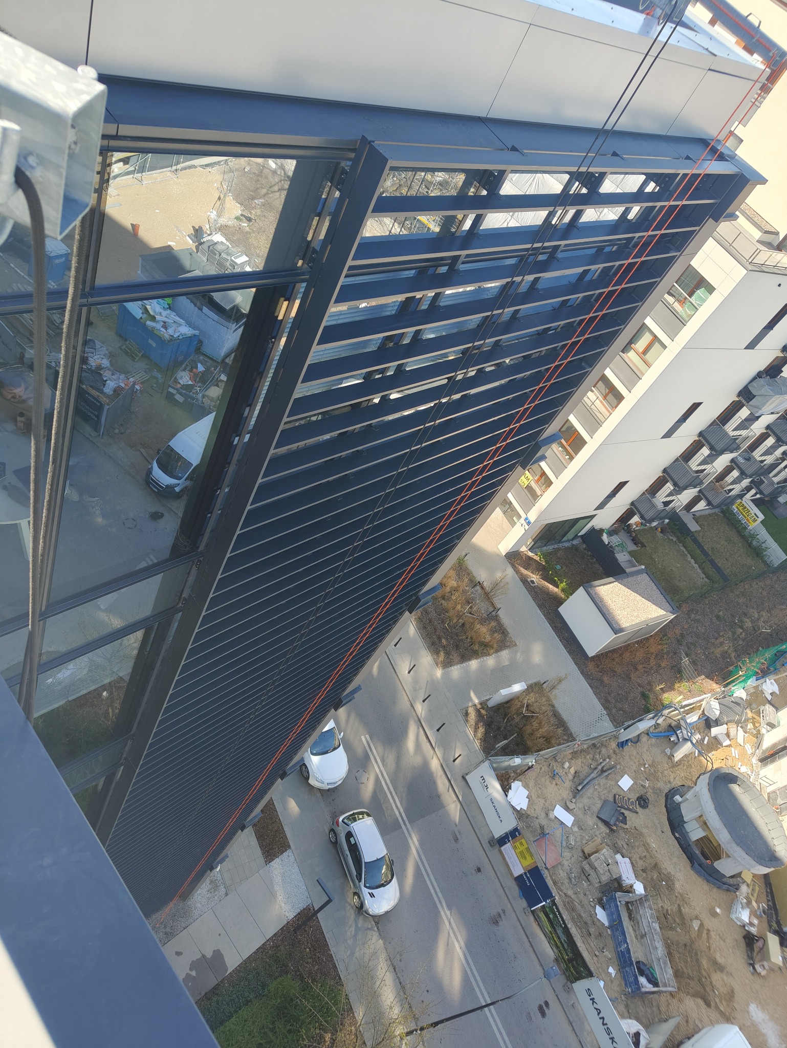 Widok z góry na nowoczesny budynek z przeszkloną fasadą i aluminiowymi żaluzjami. Na dole widoczny plac budowy, zaparkowane samochody i fragment zieleni.
