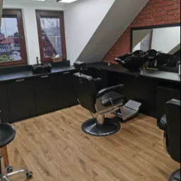 Nowoczesne wnętrze salonu fryzjerskiego z czarnymi meblami, fotelami barberskimi i podłogą z paneli. Widoczne okna i ceglana ściana, tworzące unikalny klimat.