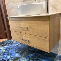 STOLARSTWO - Drewniana szafka łazienkowa z dwiema szufladami, umywalką nablatową i chromowanym kranem na tle marmurowej ściany i podłogi z niebieskiego kamienia.