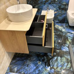 STOLARSTWO - Łazienka z umywalką nablatową na szafce z szufladami. Na podłodze i ścianach płytki imitujące kamień w odcieniach niebieskiego i szarego. Widoczny fragment toalety i bidetu.