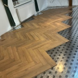 STOLARSTWO - Parkiet ułożony we wzór jodełki na podkładzie izolacyjnym, widok z góry. Fragment podłogi w trakcie montażu, jasne panele, okno z zasłonami w tle.