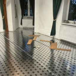 STOLARSTWO - Rozłożona folia izolacyjna pod panele podłogowe w jasnym wnętrzu z zielonymi zasłonami. Częściowo ułożone panele. Widoczny fragment rolki folii.