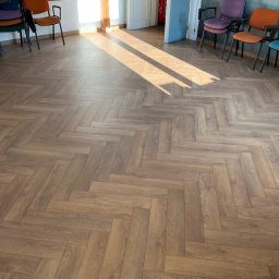 STOLARSTWO - Ułożone panele podłogowe w jodełkę, ciepłe odcienie brązu, widoczne krzesła i fragment ściany z dekoracyjnym motywem górskim. Promienie słońca padają na podłogę.