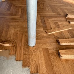STOLARSTWO - Ułożona podłoga z jasnego drewna w jodełkę wokół szarego słupa, z widocznymi stosami desek i fragmentem betonowej posadzki.