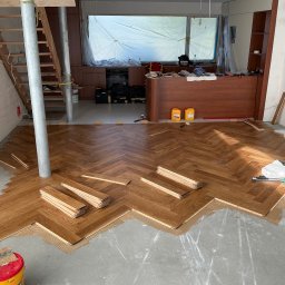 STOLARSTWO - Częściowo ułożona drewniana podłoga w jodełkę klasyczną w trakcie prac remontowych, z widocznym klejem i narzędziami na betonowym podłożu. W tle tymczasowe podpory i zakryte okno.