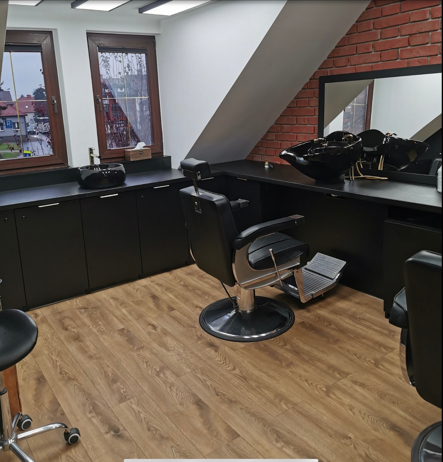 Nowoczesne wnętrze salonu fryzjerskiego z czarnymi meblami, fotelami barberskimi i podłogą z paneli. Widoczne okna i ceglana ściana, tworzące unikalny klimat.